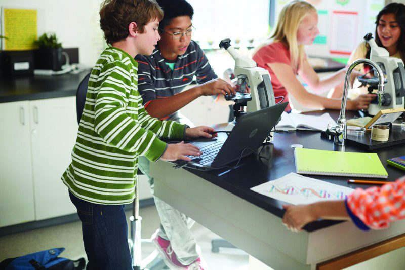 Students working with microscopes and laptops in a classroom